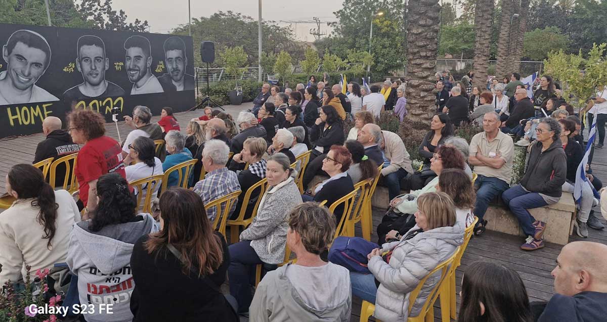פעילות למען החטופים בכפר סבא. קהל רב יושב בגינה ציבורית מול מסך עליו מוקרנים תמונות ושמות של חטופים.