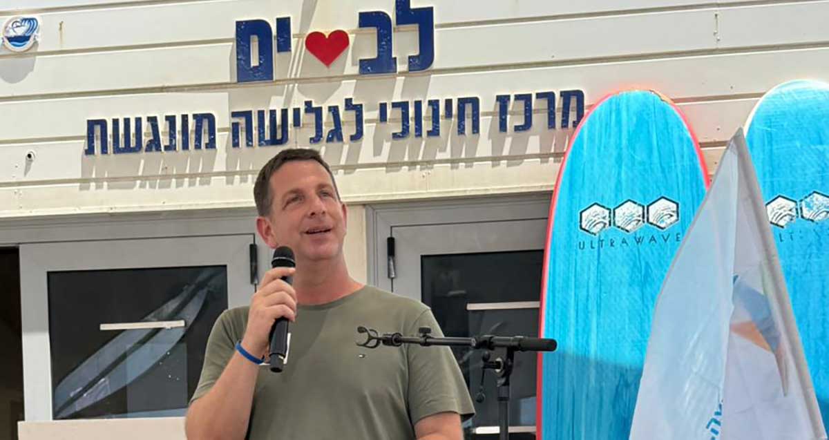 החוף השווה. ראש העיר עם מיקרופון ביד. מעליו על מבנה כיתוב: לב ים, מרכז חינוכי לגלישה מונגשת.