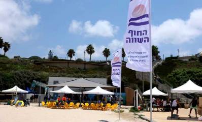 החוף השווה. צילום מכיוון הים אל החוף. במרכז בית קפה עם שמשיות לבנות. שני דגלים עם כיתוב: החוף השווה, חוף פתוח לכולם, המותאם לאנשים עם צרכים מיוחדים. ברקע הטיילת.