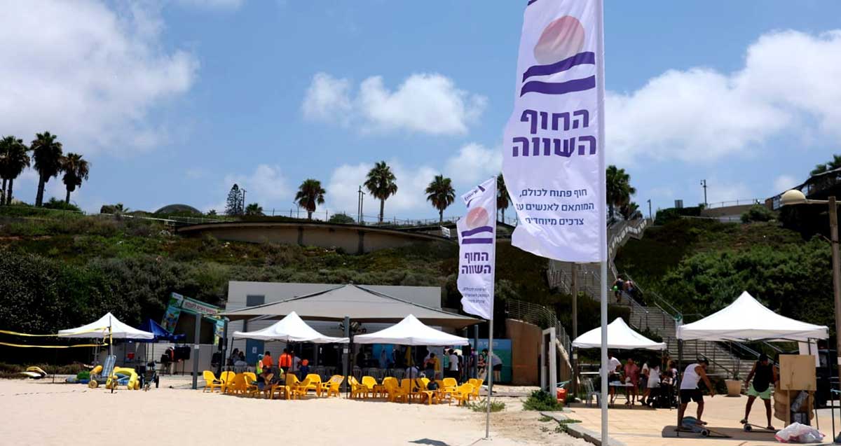 החוף השווה. צילום מכיוון הים אל החוף. במרכז בית קפה עם שמשיות לבנות. שני דגלים עם כיתוב: החוף השווה, חוף פתוח לכולם, המותאם לאנשים עם צרכים מיוחדים. ברקע הטיילת.