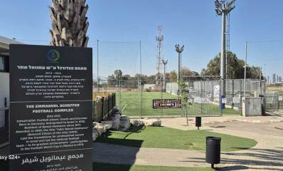 זירת השלכת הרימון במגרש הכדורגל ברמת השרון. בימין תמונה של מגרשי דשא עם עמודי תאורה, ובשמאל מבנה חד קומתי עם גדר ושלט גדול בעברית, אנגלית וערבית. הכיתוב בעברית: מתחם הכדורגל ע"ש עמנואל שפר, 1923-2012, תעשיין, שחקן, מאמן ומחנך. יליד גרמניה, עלה לארץ ב-1950, תושב רמת השרון מאז 1977 ובעל אות יקיר העיר לשנת 2010. הניח את אבני היסוד לכדורגל המקצועי בישראל והוביל את נבחרת ישראל בכדורגל לגדול הישגיה - השתתפות במשחקי גביע העולם במקסיקו 1970.