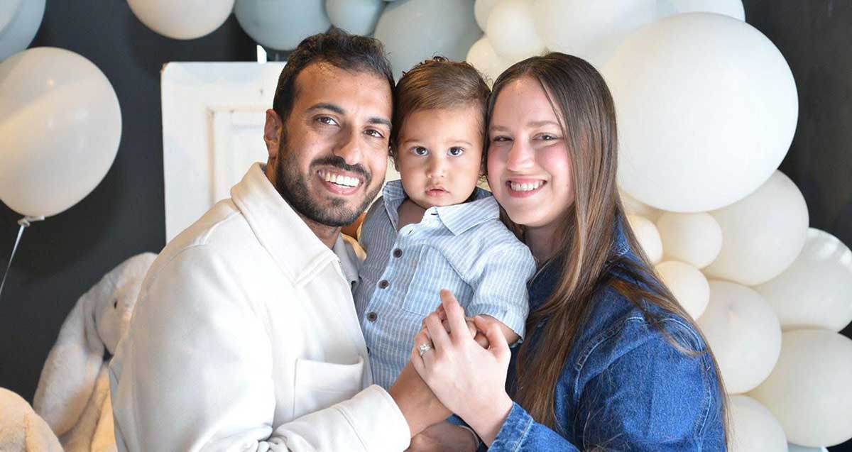 גיא ישראל עוזר ראש עיריית כפר סבא עם משפחתו, אשתו והבן על הידיים בין שניהם, ברקע בלונים לבנים ותכלת.