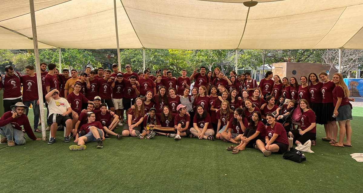 קבוצת בנים ובנות של מחנה הדר מכפר סבא. עומדים ויושבים על דשא סינטטי מתחת לצאליה