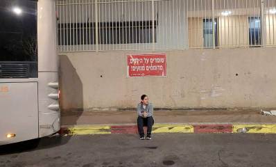 קרן קירשנר סמואל על רקע בית הספר דבורה עומר ברמת השרון. קרן יושבת על המדרכה ולידה אוטובוס. מעליה על קיר בית הספר שלט בכיתוב: שומרים על הילדים, מדוממים מנועים!