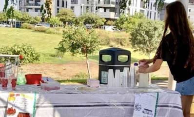 דוכן הלימונדה בפארק. על שולחן ארוך ועליו מפה ויריעת פלסטיק מונחות כוסות פלסטיק ומיכל לימונדה אותו אוחזת בחורה. בחזית השולחן שני שלטים. בצד שמאל עגלת סופרמרקט. ברקע גינה ובתי מגורים.