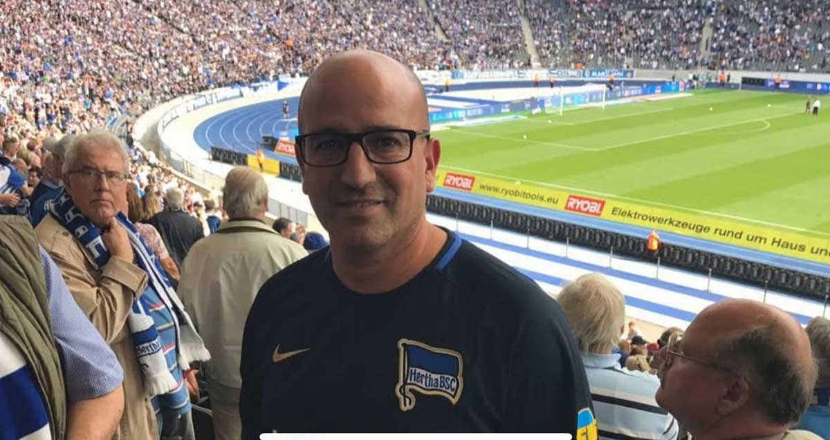 ליאור סילוק עומד בין הקהל הממלא את היציעים במגרש כדורגל. לובש חולצה כחולה עם כיתוב באנגלית: Hertha BSC