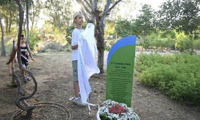 ההנצחה לזכרו של מארק קונונוביץ ז"ל. שלט גבוה בצבע ירוק עם שמו של מארק, תאריכים 1988-2024 וכיתוב רב. השלט בגינה ציבורית, ולידה ילד שהסיר את הלוט הלבן.