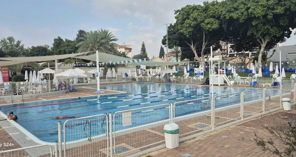 בריכת שרונית ברמת השרון. בריכה לא מקורה. ליד הבריכה משטחים עם כיסאות ומעל יריעת בד לצל.