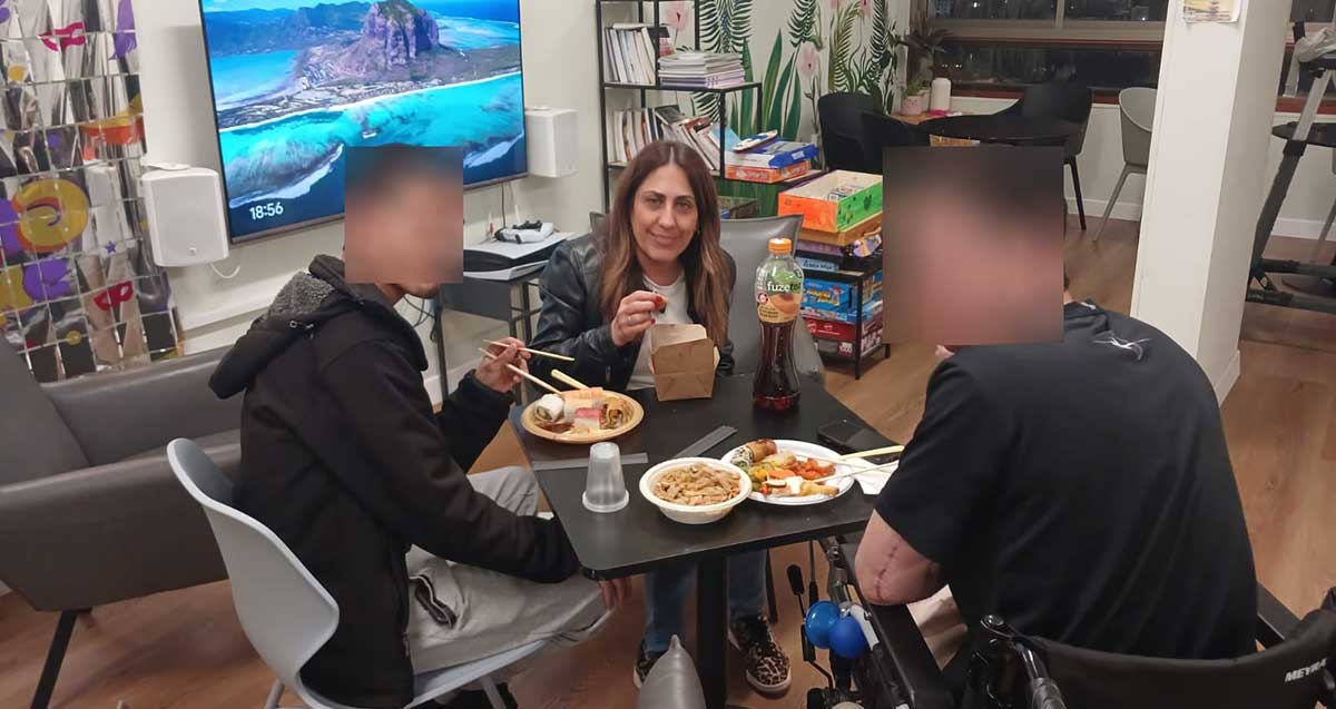 שטרית ושני לוחמים יושבים ליד שולחן עם אוכל בצלחות חד פעמיות ואוחזים שני מקלות לאכילה.