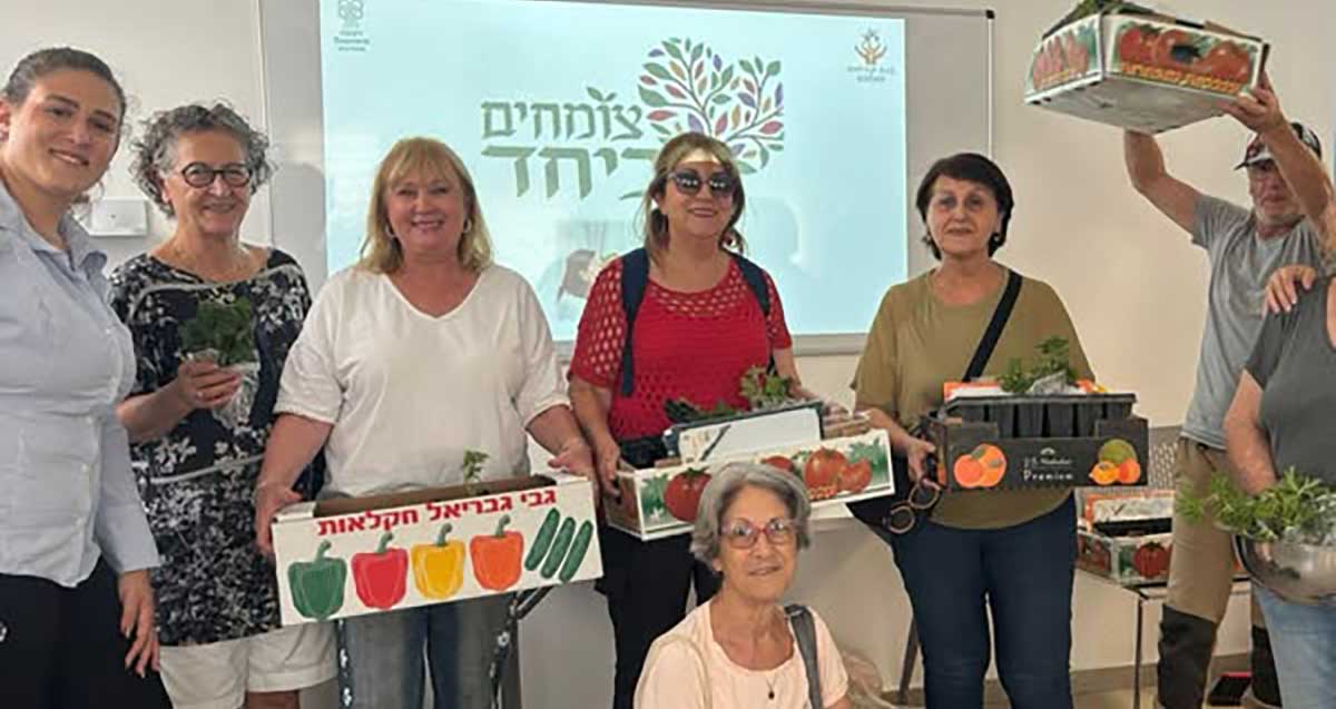 מתנדבי פרויקט "צומחים ביחד" עם ארגזים ובהם שתילים. מאחוריהם על מסך תמונה של צמח וכיתוב: צומחים ביחד.