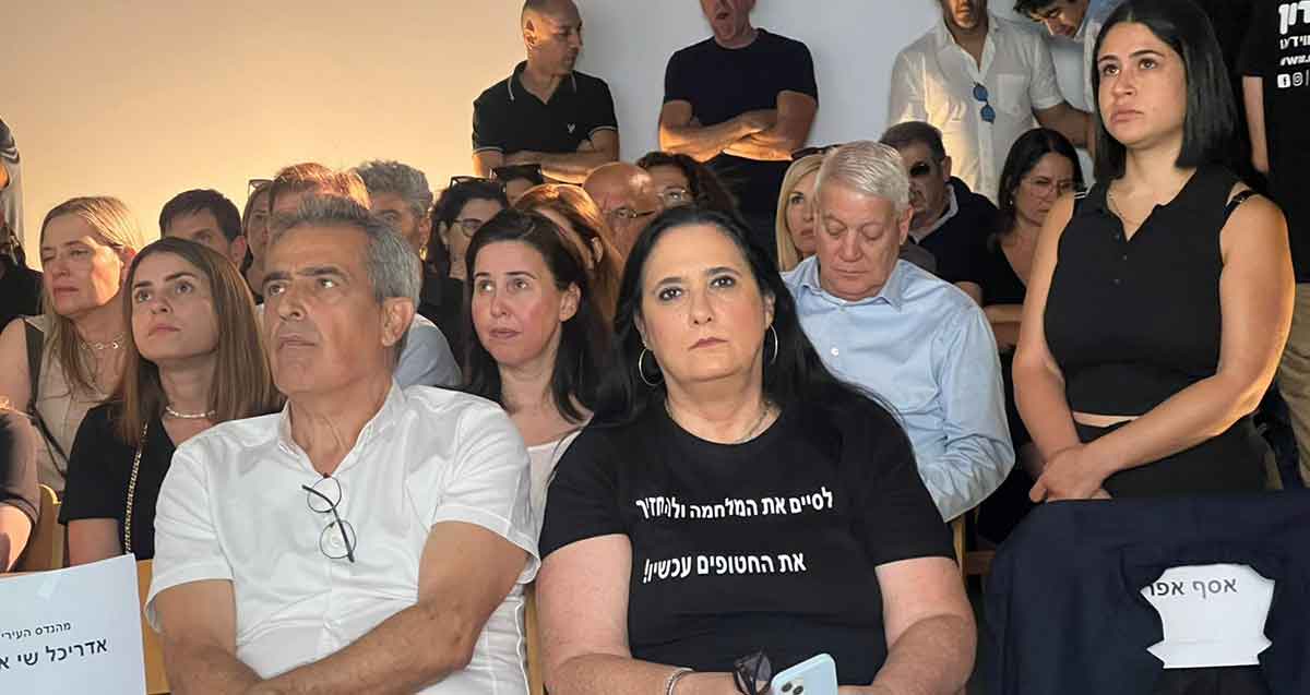 הדר שוויצר בחולצה שחורה יושבת בקהל. על חולצתה הכיתוב: לסיים את המלחמה ולהחזיר את החטופים עכשיו!