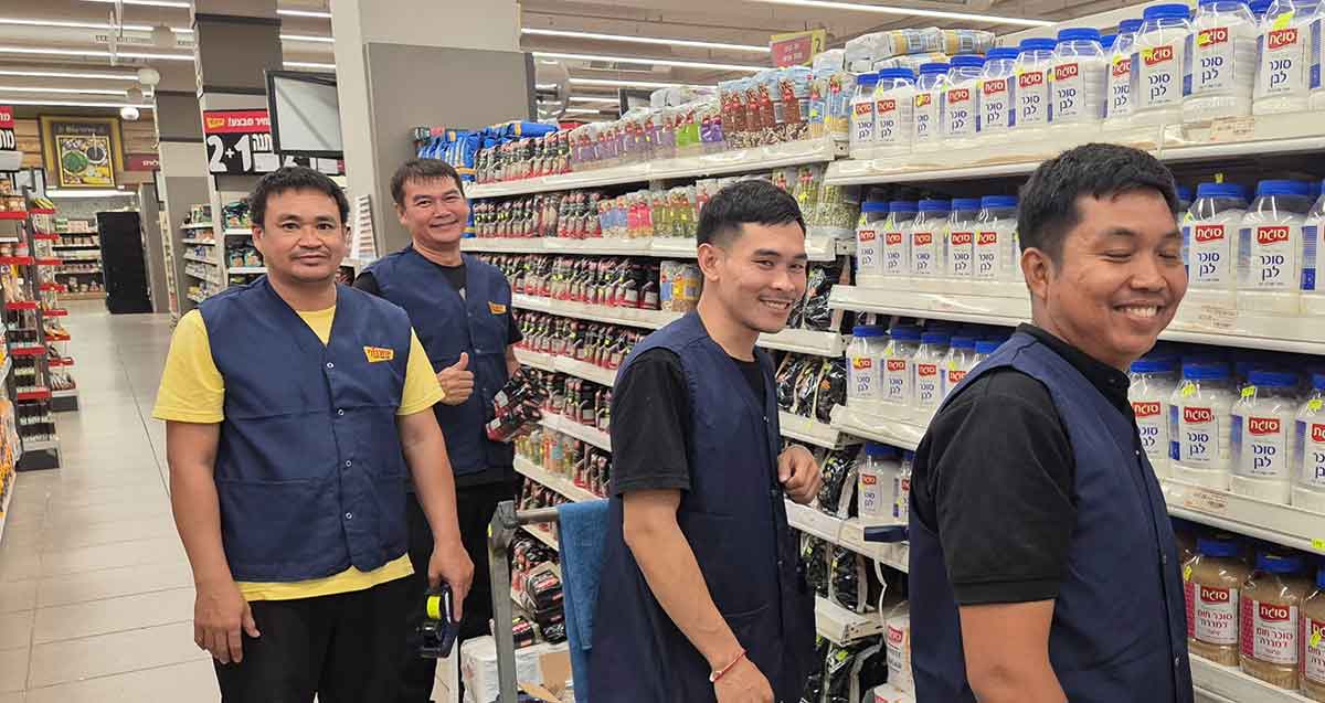 עובדים תאילנדים בסניף יוחננוף רמת השרון. ארבעה עובדים עם בגד של יוחננוף ליד מדף של מצרכים.
