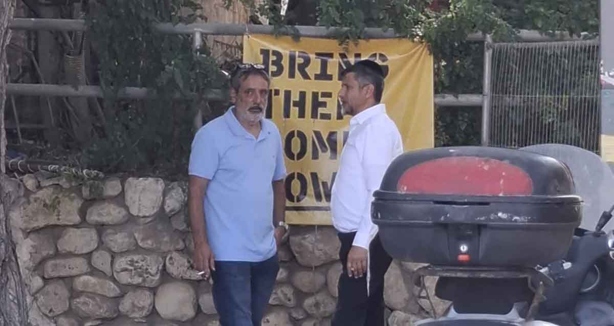 עמוס מגוז מסתודד עם זכי טיירי. ברקע שלט צהוב באנגלית: BRING THEM HOME NOW