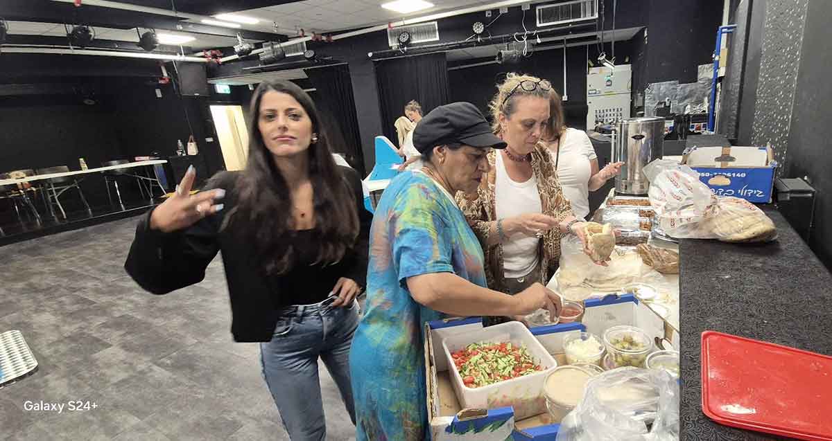 כיבוד בקלפי העובדים ברמת השרון. שולחן עם קופסאות פלסטיק עם מזון, ועובדות ממלאות פיתות. עובדת אחת עומדת מול הצלם.