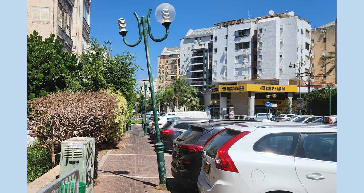 עמוד תאורה עקום בפינת הרחובות הדרור והבנים בהוד השרון. נטוע על מדרכה ליד חניית רכבים