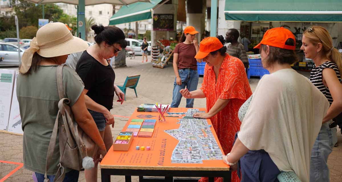 כשש נים תושבות הוד השרון מתבוננות בתוכניות עתידיות המוצבות על שולחן. השולחן והושבות נמצאות ברחוב