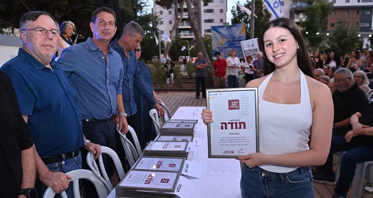 הוקרה למתנדבות ומתנדבים בהרצליה. מתנדבת עם תעודה ולידה שולחן עם תעודות וליד השולחן ראש העיר ומכובדים.