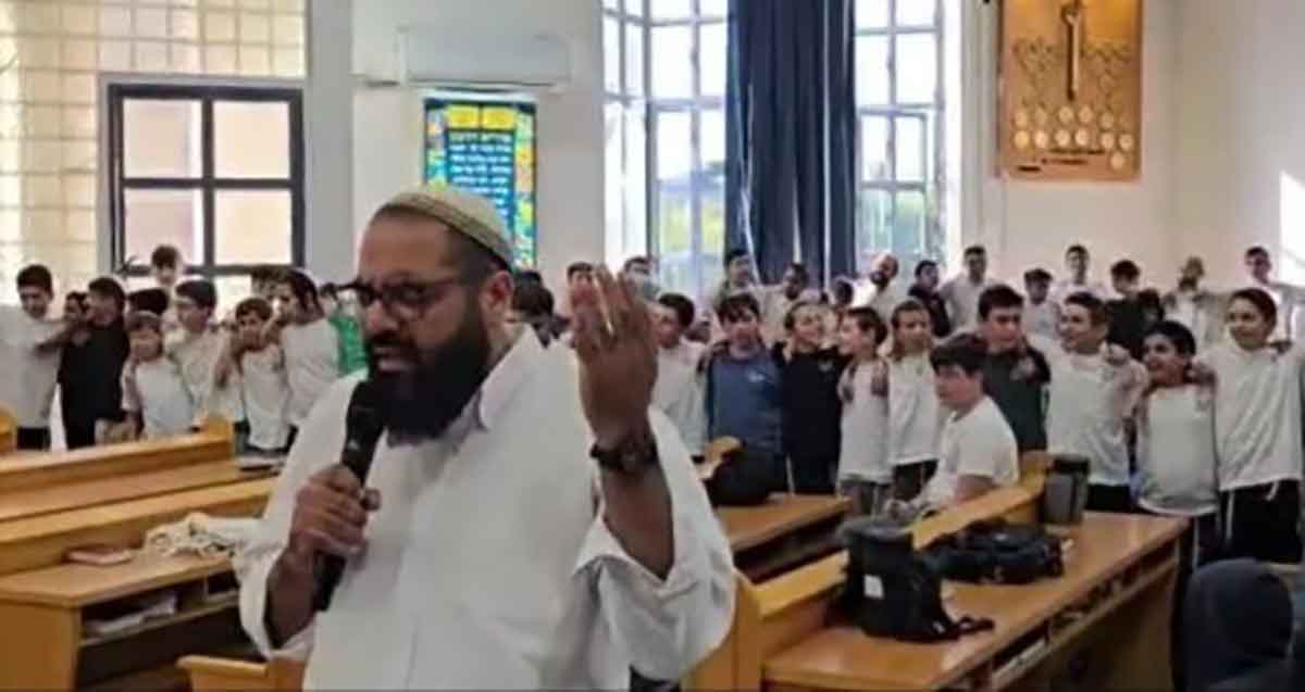 ישיבת בני עקיבא רעננה. מורה עומד עם מיקרופון ואחריו עומדים תלמידים בשורות.
