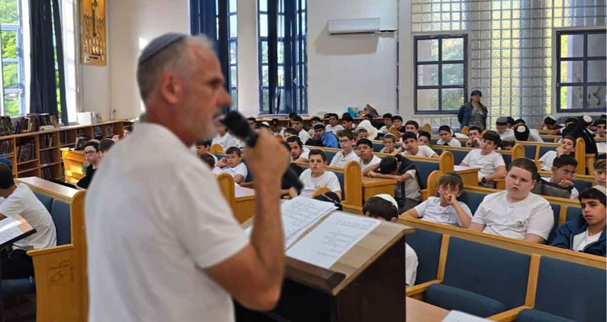 ישיבת בני עקיבא רעננה. מורה עם מיקרופון עומד ליד דוכן, לפני אולם מלא בתלמידים היושבים בשורות בספסלים.
