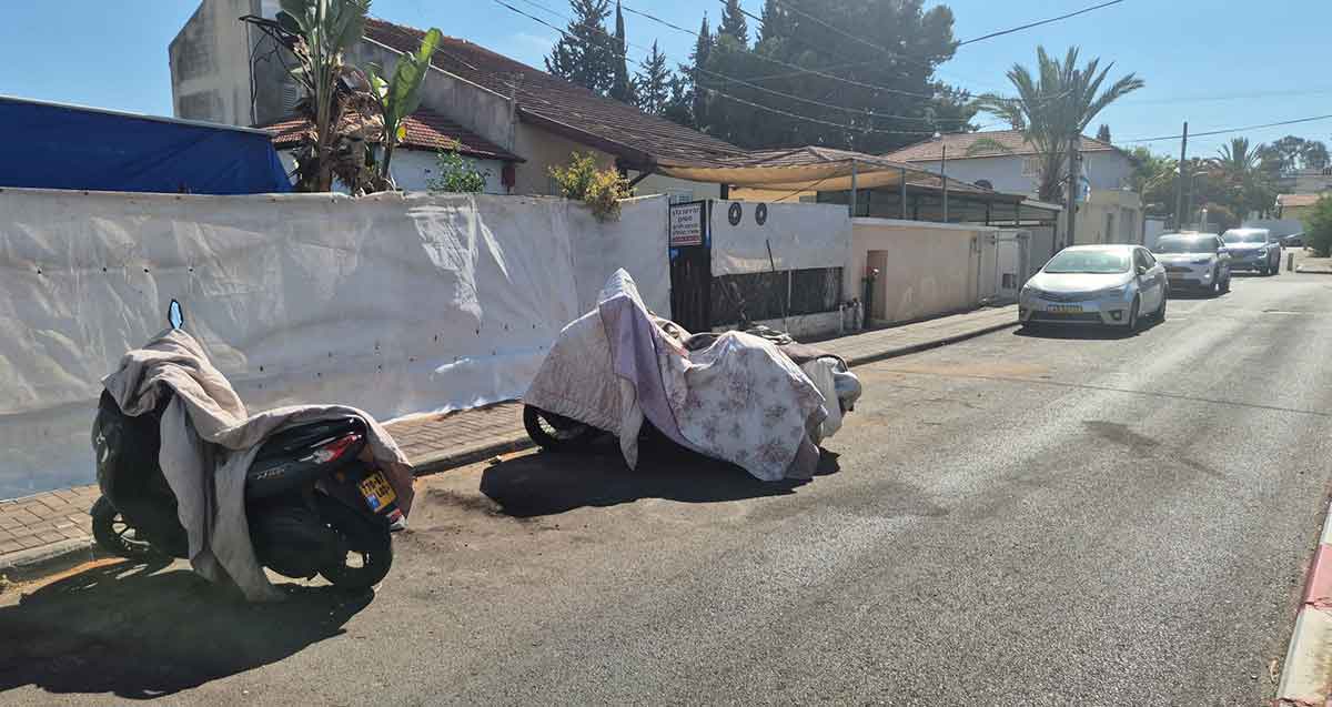מוסך ללא היתר בשכונת נווה עמל. המוסך בתוך חצר של בית והוא מוסתר ביריעה לבנה לאורך הגדר של הבניין. שני אופנועים חונים ברחוב ועליהם שמיכות.