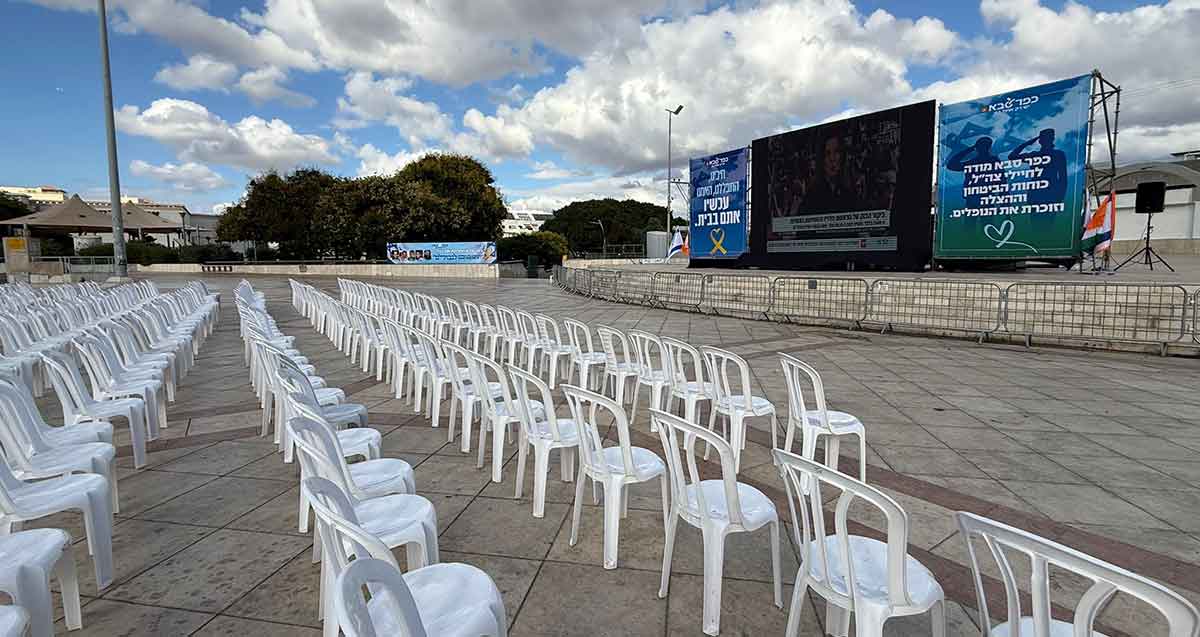 הקרנה פומבית של חזרת החטופים. מסך ענק ובשני צדדיו שלטים, מימים כיתוב: כפר סבא מודה לחיילי צה"ל, כוחות הביטחון וההצלה וזוכרת את הנופלים. משמאל הכיתוב: חיכינו, התפללנו, האמנו עכשיו אתם בבית. לפני המסך רחבה עם שורות של כיסאות פלסטיק לבנות.