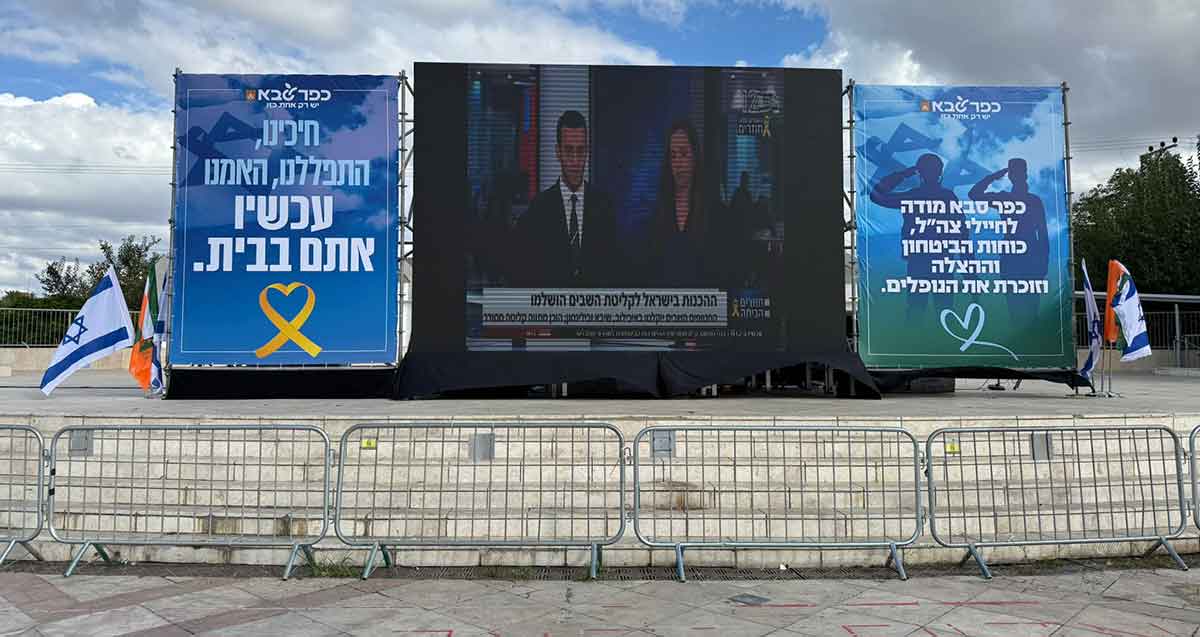 הקרנה פומבית של חזרת החטופים. מסך ענק ובשני צדדיו שלטים, מימים כיתוב: כפר סבא מודה לחיילי צה"ל, כוחות הביטחון וההצלה וזוכרת את הנופלים. משמאל הכיתוב: חיכינו, התפללנו, האמנו עכשיו אתם בבית.