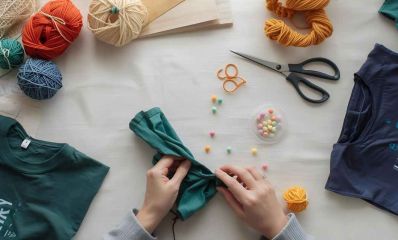 תמונה של שולחן יצירה עם ידיים גוזרות חולצות ישנות והופכות אותן לתיקי בד צבעוניים. מסביב חומרי יצירה פשוטים, גזרים, חוטים ומספריים. הדמיית בינה מלאכותית: השרון אונליין