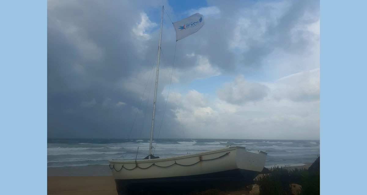 סירה עם דגל מתנוסס בראש התורן של עיריית הרצליה על החוף ליד הים הסוער והשמיים המעוננים בחוף זבולון