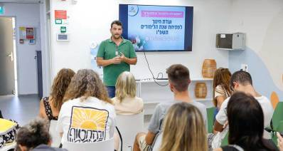 אמיר כוכבי עומד לפני קבוצה של צעירים יושבים ומאחוריו מוקרן הכיתוב: ועדת חינוך לפתיחת שנת הלימודים תשפ"ו.