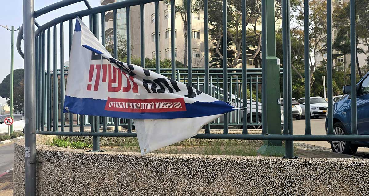 שלט להשבת החטופים מתנופף ברוח. השלט תלוי על גדר בארבע פינות, פינה אחת ניתקה.