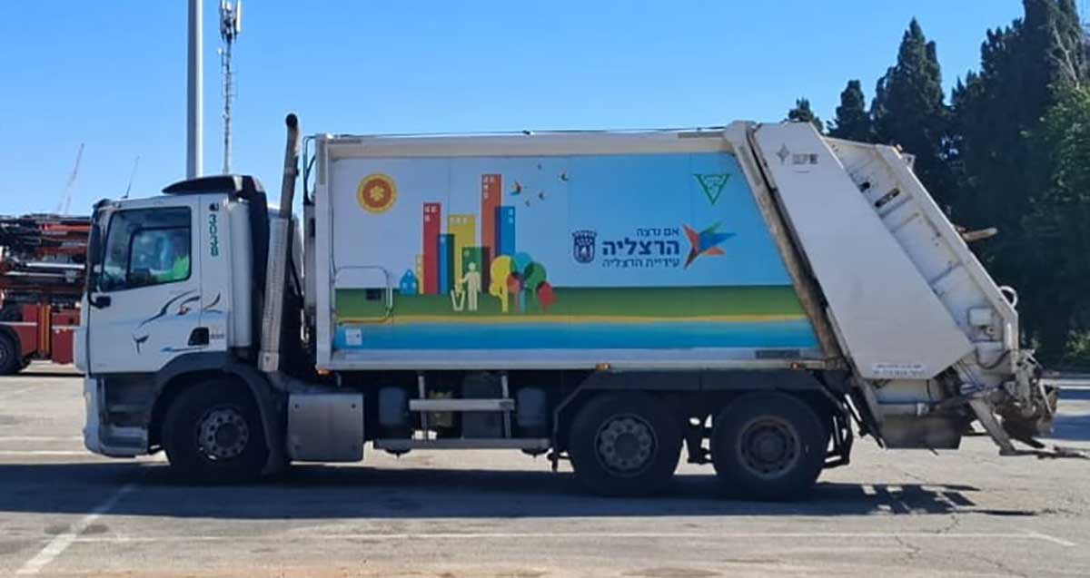 משאית פינוי אשפה של הרצליה. על דופן המשאית כיתוב: אם נרצה, הרצליה, עיריית הרצליה וסמל של העירייה. וציור על דופן המשאית של בתים ועצים, ואדם משליך אשפה לפח.