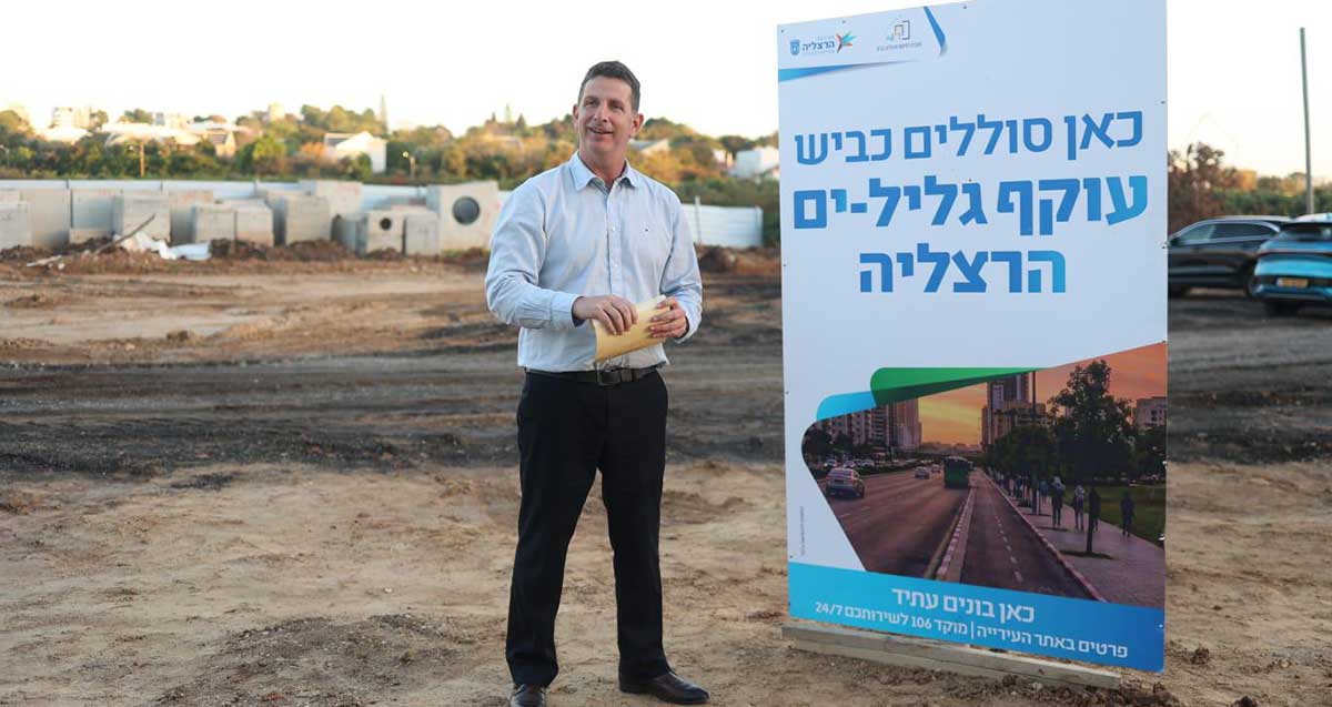 יריב פישר בשטח אדמה ובנייה כבוש בגליל ים ליד שלט רול אפ ענק שבו כתוב: כאן סוללים כביש עוקף גליל-ים הרצליה. כאן בונים עתיד