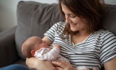 אמא צעירה יושבת על ספה, מחזיקה ומחייכת אל תינוקת רכה שישנה בזרועותיה, הדמיית בינה מלאכותית: שרון אונליין