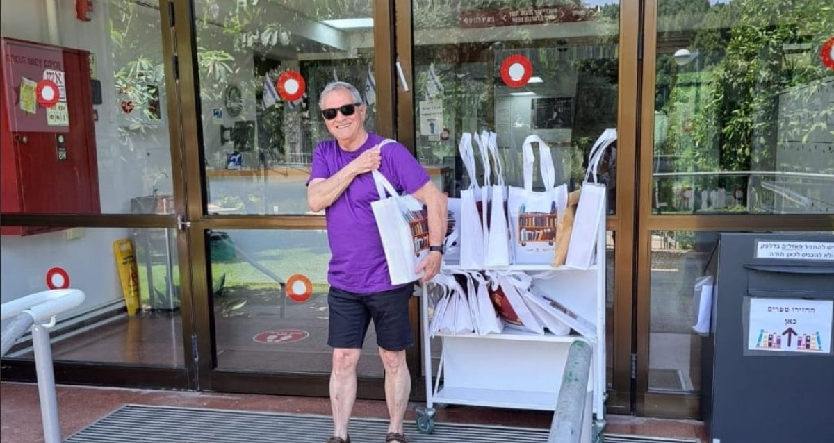 איש ע חולצה סגולה נושא על הכתף תיק ספרייה, מאחורי עגלה עם שקיות וספרים, צילום: עיריית כפר סבא