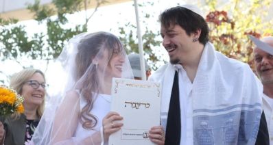 כלה עם הינומה מחזיקה את הכתבה, וחתן עם טלית עומדים מתח לחופה, צסתכלים זה על זהו ומחייכים, צילום: עיריית הרצליה