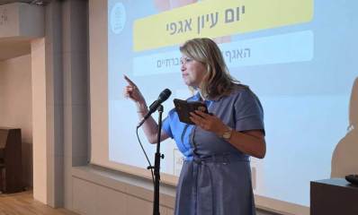 חברת מועצת העיר רעננה, זהבה לנדמן, מסיעת הרוב הדמוקרטי נואמת מעל במה. במסך מאחוריה מוקרן הכיתוב: יום עיון אגפי
