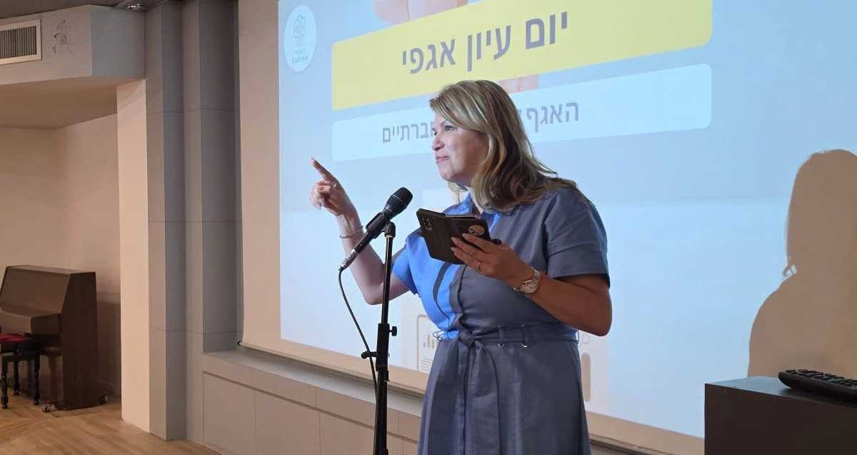 חברת מועצת העיר רעננה, זהבה לנדמן, מסיעת הרוב הדמוקרטי נואמת מעל במה. במסך מאחוריה מוקרן הכיתוב: יום עיון אגפי