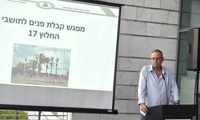 איציק רוכברגר מקבל פני תושבים חדשים בעיר. נואם מעל פודיום ולידו מסך עליו מוקרן: מפגש קבלת פנים לתושבי החלוץ 17