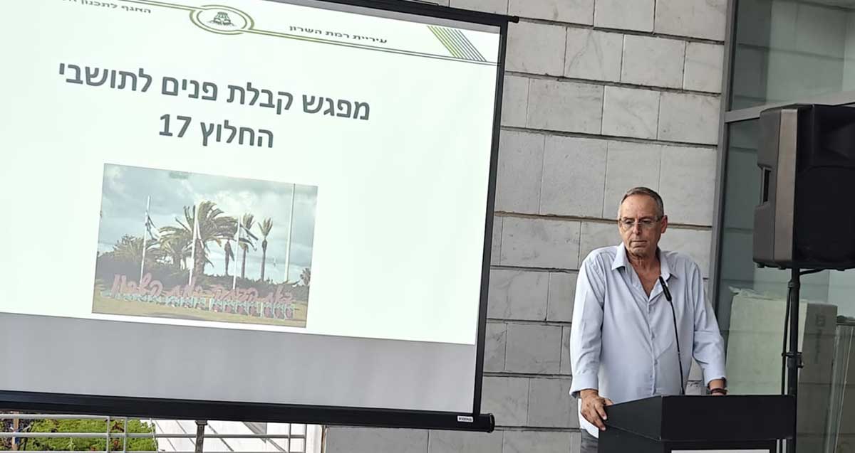 איציק רוכברגר מקבל פני תושבים חדשים בעיר. נואם מעל פודיום ולידו מסך עליו מוקרן: מפגש קבלת פנים לתושבי החלוץ 17