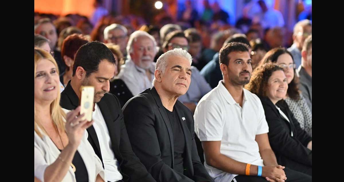 גיא שראל עם יאיר לפיד יושבים באולם עם שורות של קהל.