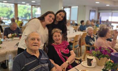 תלמידי אורט כפר סבא עם שורדי שואה. שתי תלמידות עומדות מאחורי זוג שורדי שואה היושבים ליד שולחן. מסביבם יושבים שורדי שואה ליד שולחנות.
