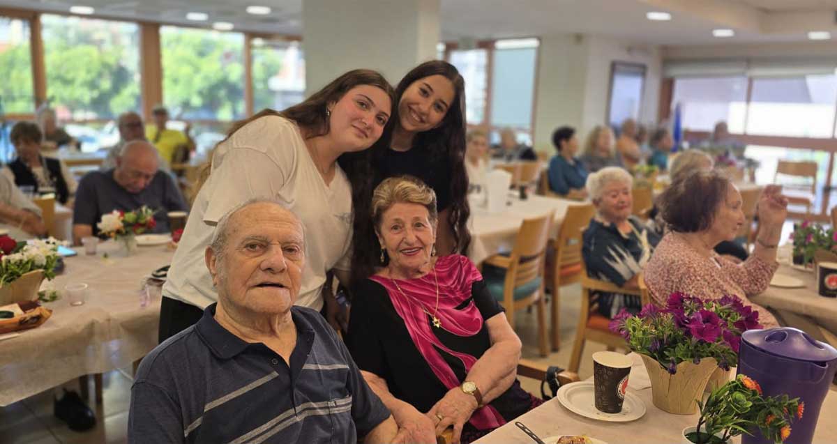 תלמידי אורט כפר סבא עם שורדי שואה. שתי תלמידות עומדות מאחורי זוג שורדי שואה היושבים ליד שולחן. מסביבם יושבים שורדי שואה ליד שולחנות.