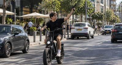 צעיר עם אקדח רוכב על אופניים חשמליים. איור AI. הצעיר רוכב בכביש ואוחז את האופניים ביד אחת והיד השנייה כלפי מעלה והצידה עם אקדח.