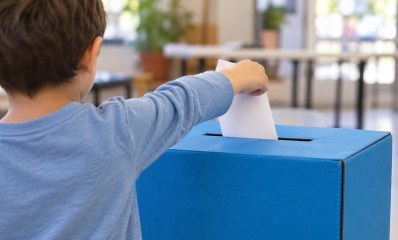 אילוסטרציה שנוצרה בבינה מלאכותית של ילד מצולם מאחור מכניס פתק לקלפי כחולה